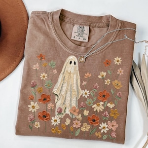 May include: A brown short-sleeve t-shirt featuring an embroidered ghost surrounded by a variety of colorful wildflowers in shades of orange, yellow, pink, and green. The shirt has a Comfort Colors brand label inside the collar and a crew neckline.