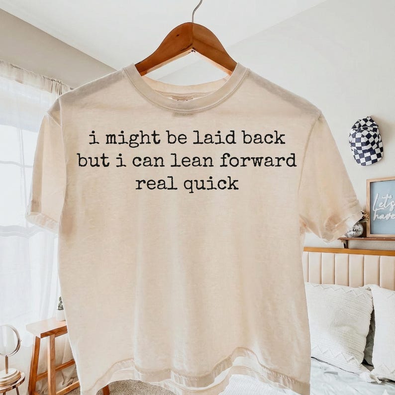May include: A cream-colored short-sleeve t-shirt hanging on a wooden hanger. The shirt features black typewriter-style text printed on the front that reads: I might be laid back but i can lean forward real quick. The fabric has a subtle textured appearance.