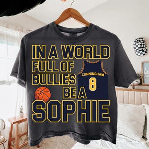 May include: A charcoal gray short-sleeve t-shirt featuring the yellow text IN A WORLD FULL OF BULLIES BE A SOPHIE. The design includes a graphic of an orange basketball and a navy blue basketball jersey with the name CUNNINGHAM and number 8.
