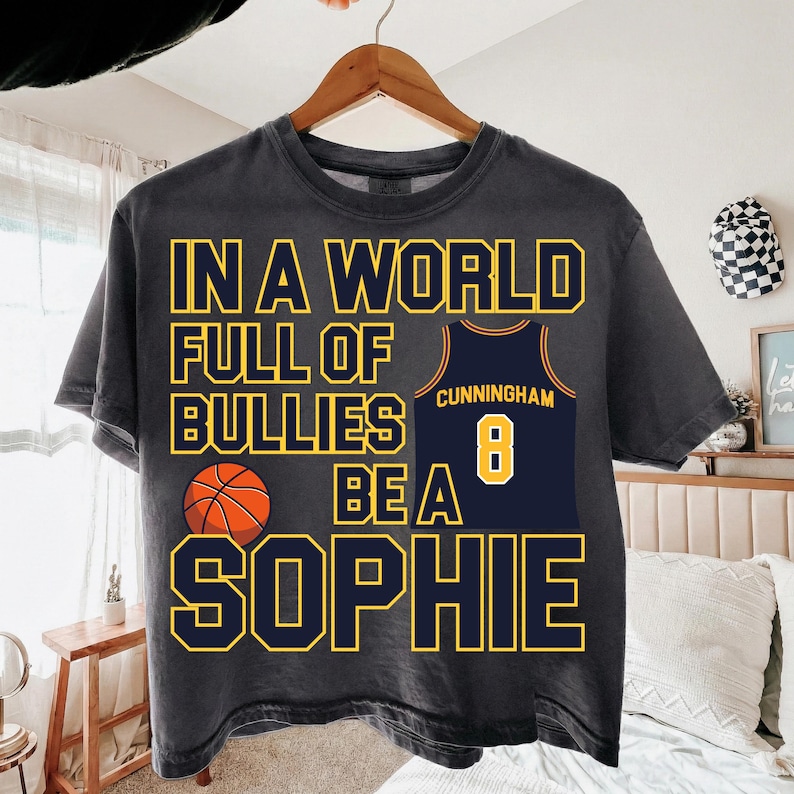 May include: A charcoal gray short-sleeve t-shirt featuring the yellow text IN A WORLD FULL OF BULLIES BE A SOPHIE. The design includes a graphic of an orange basketball and a navy blue basketball jersey with the name CUNNINGHAM and number 8.