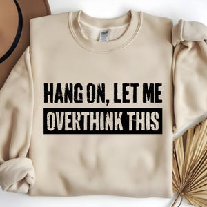 Op de afbeelding: Een beige sweatshirt met de tekst "HANG ON, LET ME OVERTHINK THIS" in zwarte blokletters. De tekst is gecentreerd op de voorkant van de sweatshirt, met de woorden "OVERTHINK THIS" in een zwarte rechthoek.