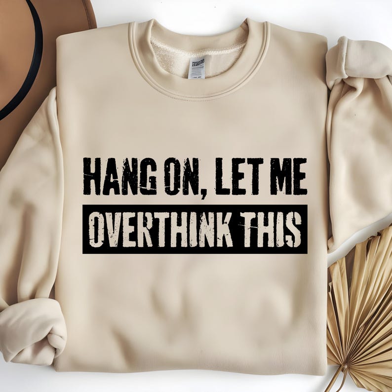 Op de afbeelding: Een beige sweatshirt met de tekst "HANG ON, LET ME OVERTHINK THIS" in zwarte blokletters. De tekst is gecentreerd op de voorkant van de sweatshirt, met de woorden "OVERTHINK THIS" in een zwarte rechthoek.