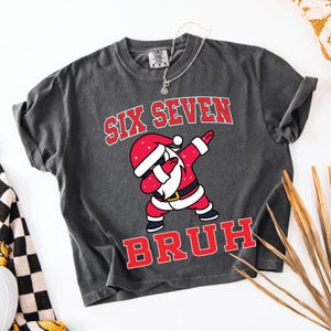 Könnte beinhalten: Dunkelgraues T-Shirt mit dem Text "SIX SEVEN BRUH" in roten Buchstaben. Eine Cartoon-Figur des Weihnachtsmanns in rotem Anzug und Hut dabbt. Eine silberne Halskette ist über dem Shirt drapiert.