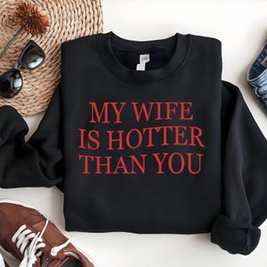 May include: Black crew neck sweatshirt with the red text "MY WIFE IS HOTTER THAN YOU". The sweatshirt is displayed on a white surface with sunglasses, a woven basket, and brown trainers.