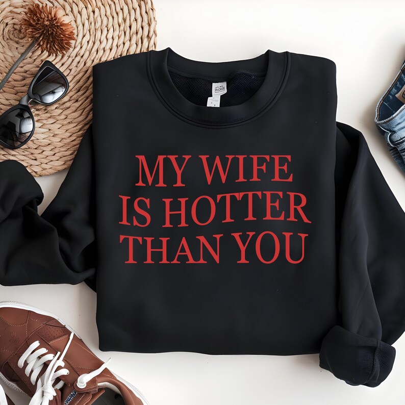 May include: Black crew neck sweatshirt with the red text "MY WIFE IS HOTTER THAN YOU". The sweatshirt is displayed on a white surface with sunglasses, a woven basket, and brown trainers.