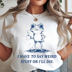 Op de afbeelding: Wit T-shirt met een blauwe illustratie van een kikker die een koffiemok vasthoudt. De kikker staat op een grasveld. De tekst "I HAVE TO SAY WEIRD STUFF OR I'LL DIE" staat eronder in het blauw.