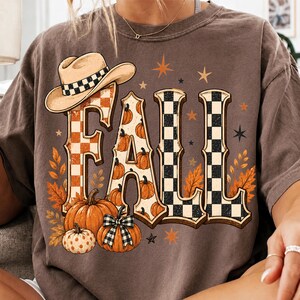 Puede incluir: Una camiseta marrón de manga corta con un estampado gráfico de la palabra FALL en letras grandes. Las letras están decoradas con calabazas naranjas, un sombrero de vaquero y un patrón de cuadros blancos y negros. Hojas de otoño y estrellas rodean el texto.