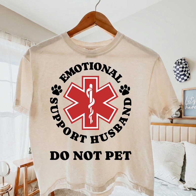May include: A beige t-shirt with a circular design featuring a red medical symbol and the words "EMOTIONAL SUPPORT HUSBAND DO NOT PET" in black. The design includes paw prints and a medical staff symbol.