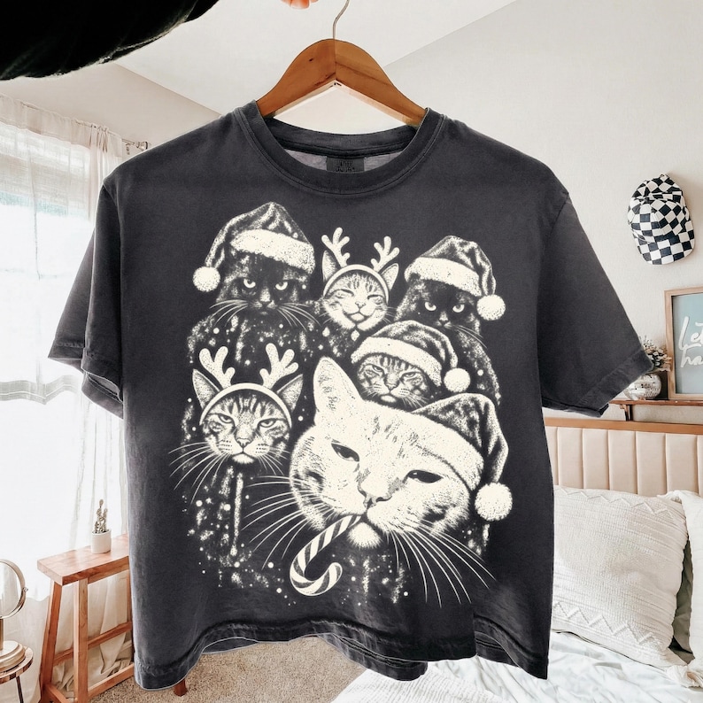 Puede incluir: Una camiseta gris carb&oacute;n con un estampado gr&aacute;fico blanco de varios gatos con gorros de Pap&aacute; Noel y astas de reno. Un gato en primer plano sostiene un bast&oacute;n de caramelo en la boca. El dise&ntilde;o incluye peque&ntilde;as motas blancas como estrellas.