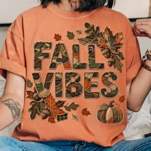 May include: A burnt orange short-sleeve t-shirt featuring the text FALL VIBES in a camouflage and plaid patchwork pattern. The design is surrounded by various autumn leaves, acorns, and a pumpkin, all rendered in a stitched patchwork style.
