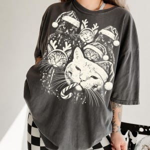 Puede incluir: Una camiseta gris carb&oacute;n de manga corta con un estampado gr&aacute;fico blanco de varios gatos con gorros de Pap&aacute; Noel y astas de reno. Un gato central sostiene un bast&oacute;n de caramelo en la boca, rodeado por un patr&oacute;n festivo de nieve.