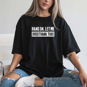 Op de afbeelding: Zwart T-shirt met de witte tekst "HANG ON, LET ME OVERTHINK THIS". Het shirt heeft een relaxte pasvorm, geschikt voor casual wear. Het model draagt een blauwe jeans.