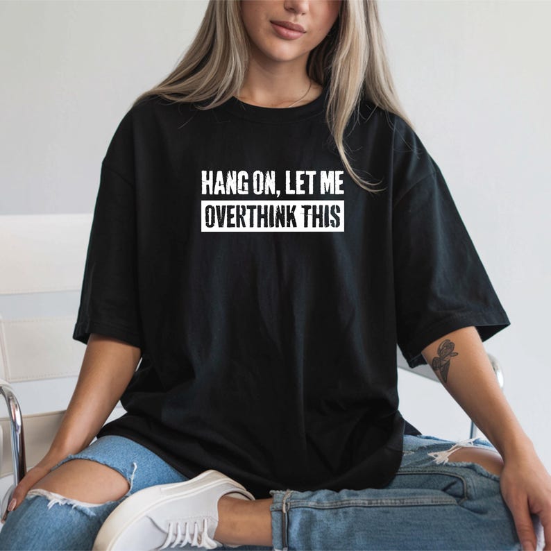 Op de afbeelding: Zwart T-shirt met de witte tekst "HANG ON, LET ME OVERTHINK THIS". Het shirt heeft een relaxte pasvorm, geschikt voor casual wear. Het model draagt een blauwe jeans.