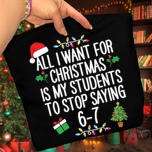 May include: Black pillow with the text "ALL I WANT FOR CHRISTMAS IS MY STUDENTS TO STOP SAYING 6-7". The design includes a Santa hat, holly, a Christmas tree, a gift, and string lights.