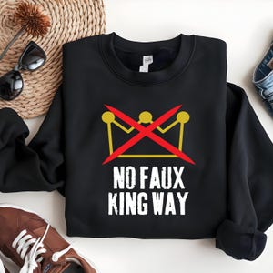 Peut inclure: Sweat-shirt noir avec un motif graphique. Le motif présente une couronne jaune barrée d'une ligne rouge et le texte "NO FAUX KING WAY" en blanc. Le sweat-shirt est de style col rond.
