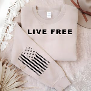 May include: A beige sweatshirt with the words "LIVE FREE" in bold black letters. The left sleeve features a distressed American flag design in black and white. The sweatshirt is folded on a patterned surface.