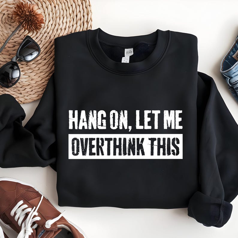 Op de afbeelding: Zwarte sweatshirt met de witte tekst "HANG ON, LET ME OVERTHINK THIS". De tekst is in hoofdletters. De sweatshirt heeft een ronde hals en lijkt van zacht materiaal te zijn.