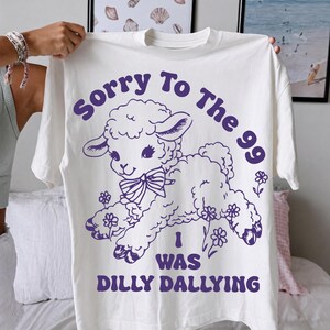May include: A white short-sleeved t-shirt featuring a purple graphic of a lamb wearing a bow. The text reads "Sorry To The 99" above the lamb and "I WAS DILLY DALLYING" below it, surrounded by small flower illustrations.