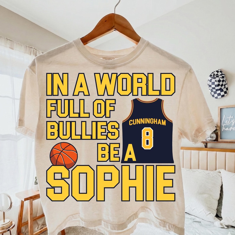 May include: A cream-colored short-sleeve t-shirt featuring the yellow text IN A WORLD FULL OF BULLIES BE A SOPHIE, a graphic of an orange basketball, and a navy blue basketball jersey with the name CUNNINGHAM and the number 8 in yellow.