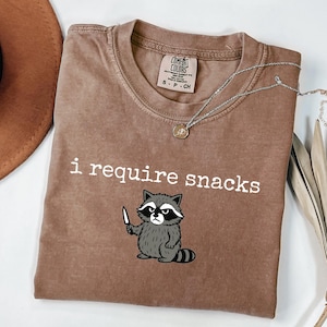 May include: A brown t-shirt with the text "i require snacks" and a cartoon raccoon holding a knife. The raccoon is gray and white with a black mask. A silver necklace with a small pendant is on the shirt.