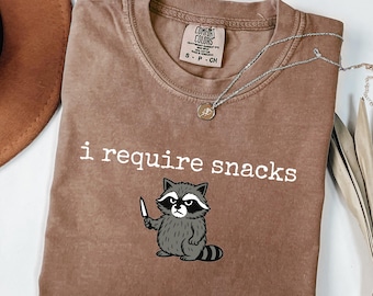 以下が含まれることがあります： 「i require snacks」の文字とナイフを持ったアライグマのイラストがプリントされた茶色のTシャツ。アライグマはグレーと白。小さなペンダントが付いたシルバーのネックレスもシャツの上にあります。