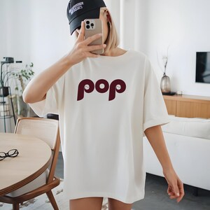 May include: Cream-colored t-shirt with the word "pop" in a burgundy, stylized font. The shirt has short sleeves and a relaxed fit. A black baseball cap is worn on the head.