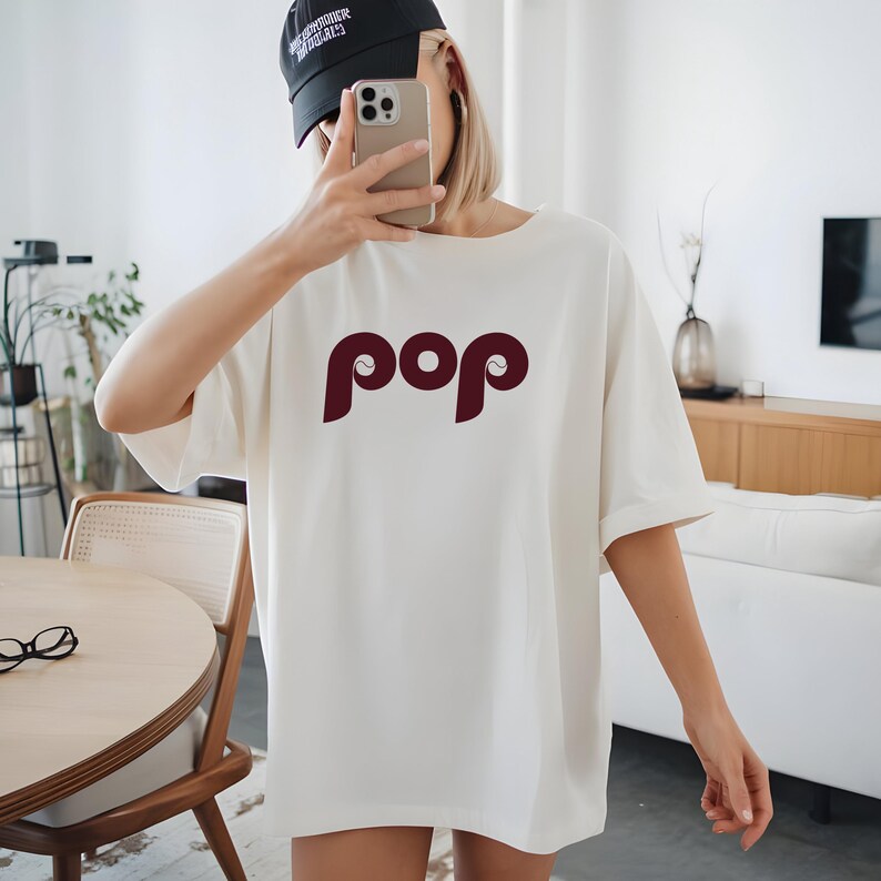 May include: Cream-colored t-shirt with the word "pop" in a burgundy, stylized font. The shirt has short sleeves and a relaxed fit. A black baseball cap is worn on the head.