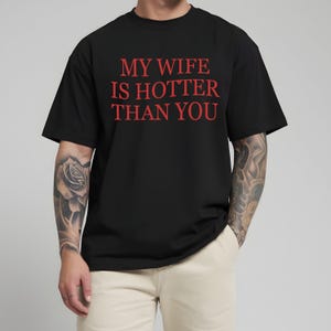 May include: Black t-shirt with the red text "MY WIFE IS HOTTER THAN YOU". The shirt has short sleeves and a crew neck. The person wearing the shirt is also wearing beige trousers.