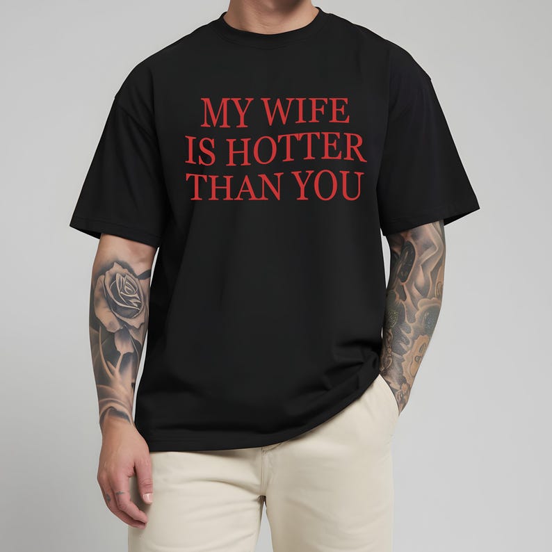 May include: Black t-shirt with the red text "MY WIFE IS HOTTER THAN YOU". The shirt has short sleeves and a crew neck. The person wearing the shirt is also wearing beige trousers.
