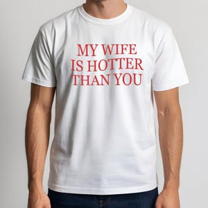 May include: White t-shirt with the red text "MY WIFE IS HOTTER THAN YOU". The shirt has short sleeves and a crew neck. The text is centered on the front of the shirt.