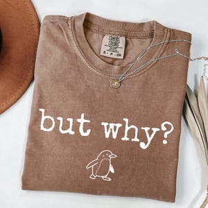 Puede incluir: Una camiseta marrón con la frase blanca "but why?" y un dibujo de un pingüino. La camiseta tiene cuello redondo y una etiqueta que dice "Comfort Colors". Un collar plateado con un colgante está sobre la camiseta.