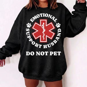 May include: Black sweatshirt with the text "EMOTIONAL SUPPORT HUSBAND DO NOT PET" in white. A red medical symbol is in the center of the design, surrounded by the text. Paw prints are on either side of the medical symbol.