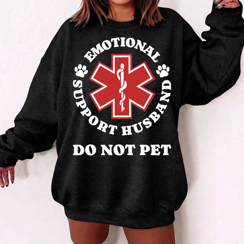 May include: Black sweatshirt with the text "EMOTIONAL SUPPORT HUSBAND DO NOT PET" in white. A red medical symbol is in the center of the design, surrounded by the text. Paw prints are on either side of the medical symbol.