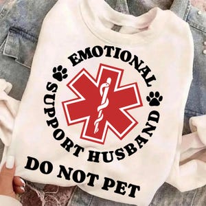 May include: White crew neck sweatshirt with the text "EMOTIONAL SUPPORT HUSBAND DO NOT PET" in black. A red cross-shaped emblem with a white staff is in the center, surrounded by the text. Two black paw prints are on either side of the emblem.