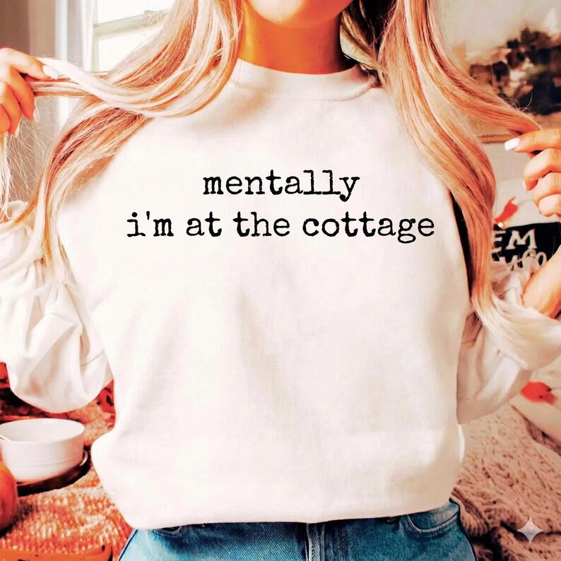 Puede incluir: Sudadera blanca con el texto negro "mentally I'm at the cottage". La sudadera es de cuello redondo. La imagen muestra a una persona que lleva la sudadera.
