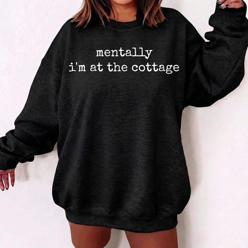 Puede incluir: Sudadera negra con el texto blanco "mentally i'm at the cottage". La sudadera de cuello redondo y manga larga es de color negro liso. El texto est&aacute; centrado en la parte delantera de la sudadera.