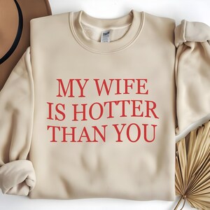 May include: A cream-coloured sweatshirt featuring the red text "MY WIFE IS HOTTER THAN YOU." The long-sleeved, crew neck top has a relaxed fit. The garment is made of a soft material.