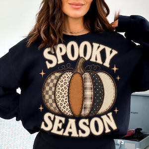 May include: A dark navy blue long-sleeve sweatshirt featuring a large central graphic of a pumpkin composed of various patterned segments including leopard print, plaid, and herringbone. The text SPOOKY SEASON is printed in a cream-colored font.