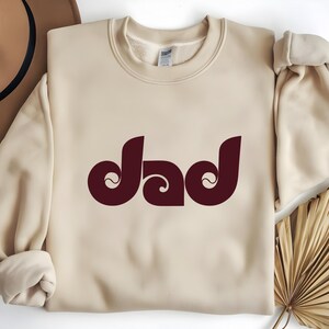 May include: A cream-colored sweatshirt with the word "dad" in a burgundy, retro-style font. The word is centered and has a unique, swirling design. The sweatshirt has a crew neck and long sleeves.