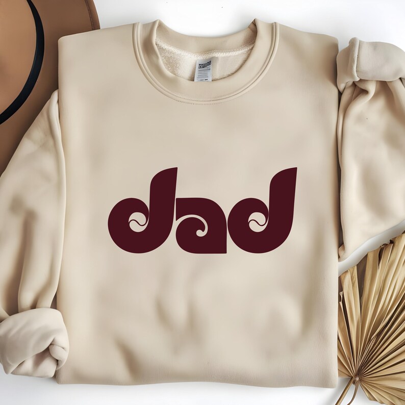 May include: A cream-colored sweatshirt with the word "dad" in a burgundy, retro-style font. The word is centered and has a unique, swirling design. The sweatshirt has a crew neck and long sleeves.