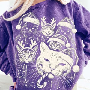 Puede incluir: Una sudadera morada con efecto lavado mineral que presenta un estampado gr&aacute;fico blanco de varios gatos con gorros de Pap&aacute; Noel y astas de reno, con un gato sosteniendo un bast&oacute;n de caramelo en la boca. El dise&ntilde;o muestra varios rostros felinos.