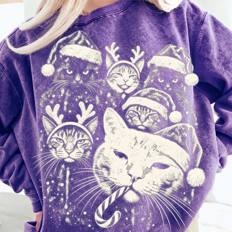 Puede incluir: Una sudadera morada con efecto lavado mineral que presenta un estampado gr&aacute;fico blanco de varios gatos con gorros de Pap&aacute; Noel y astas de reno, con un gato sosteniendo un bast&oacute;n de caramelo en la boca. El dise&ntilde;o muestra varios rostros felinos.