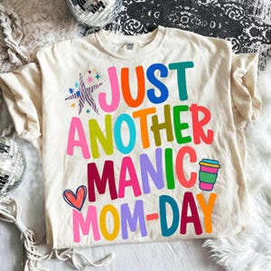 May include: Cream-colored t-shirt with the text "JUST ANOTHER MANIC MOM-DAY" in various bright colors. The shirt features a heart, a coffee cup, and star graphics. The shirt is laid on a white fluffy surface.