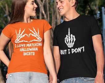 May include: Two people wearing matching Halloween t-shirts. One shirt is orange with a skeleton hand heart design and the text "I LOVE MATCHING HALLOWEEN SHIRTS". The other shirt is black with a skeleton hand giving the middle finger and the text "BUT I DON'T".