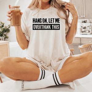 Op de afbeelding: Cr&egrave;mekleurig T-shirt met de zwarte tekst "HANG ON, LET ME OVERTHINK THIS". De persoon draagt witte sokken met zwarte strepen en houdt een glas ijskoffie vast.