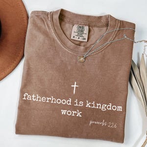 May include: A brown Comfort Colors t-shirt with the text "fatherhood is kingdom work" and a cross graphic. The shirt also features the text "proverbs 22:6". A silver necklace with a pendant is draped across the shirt.