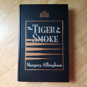 May include: A hardcover book titled "The Tiger in the Smoke" by Margery Allingham. The book has a black cover with gold lettering and accents. The top of the book reads "The Best Mysteries of All Time".