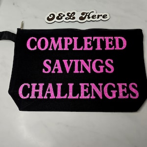 May include: Black zippered pouch with the text "COMPLETED SAVINGS CHALLENGES" in bright pink glitter lettering. The pouch has a small black handle on the side. A wooden sign that says "O&L Here" is in the background.