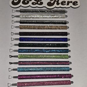 May include: A collection of twelve metal dotting tools with rhinestone-encrusted handles in various colors including silver, gold, black, blue, green, and pink, each featuring a triangular metal tip and the text J&L Here at the top.