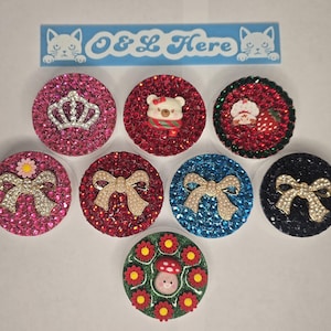 May include: Eight circular phone grips arranged in a group, each covered in colorful rhinestones. Designs include a silver crown, a bear, a strawberry, bows, and a mushroom surrounded by red flowers. A blue label at the top reads O & L Here.